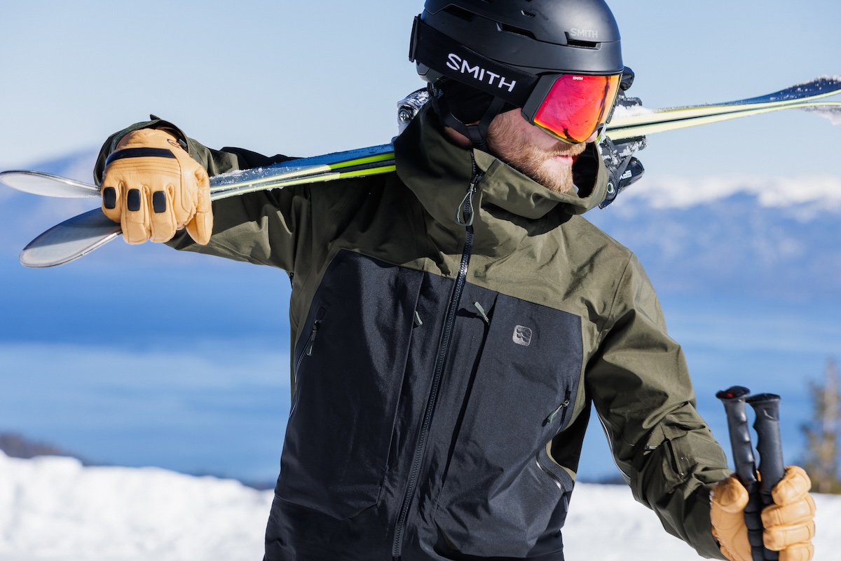 A man wearing Smith goggles looks off into the distance while carrying skis on his shoulder