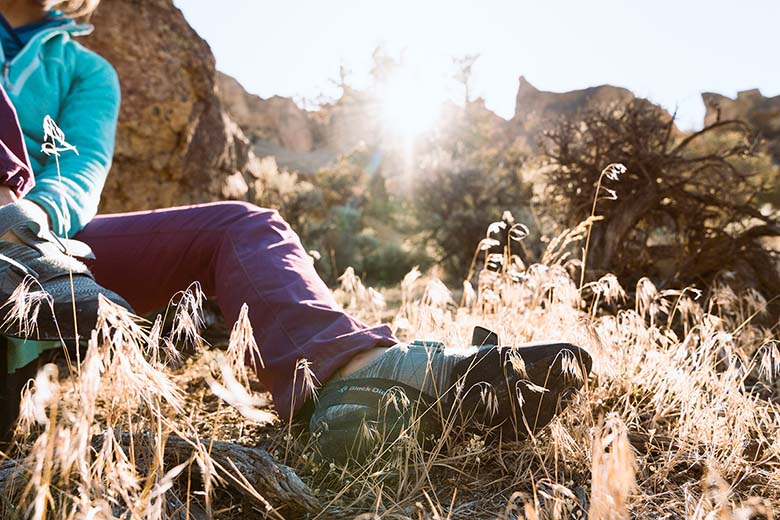 A woman puts on a climbing shoe while sitting on the ground in bright, late day sunlight