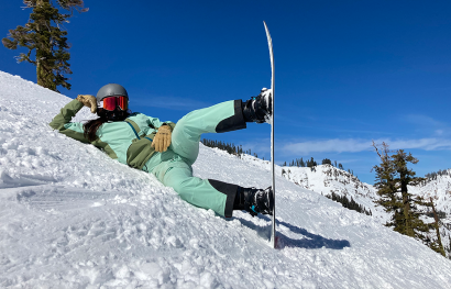 A woman lounging on the side of a snowy bank with a snowboard. 