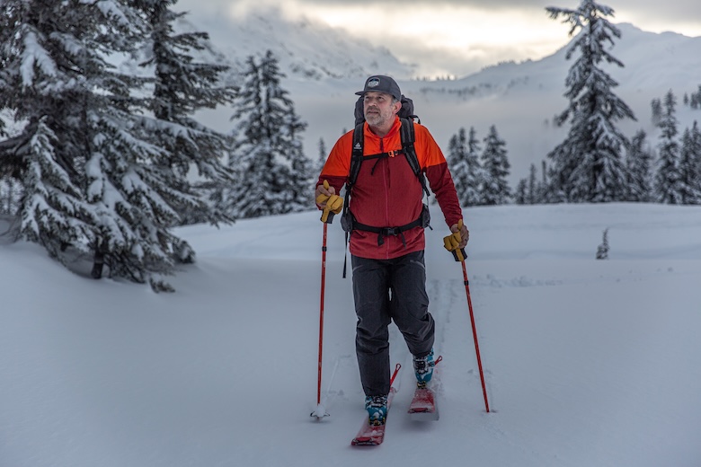 A man in a red jacket skins up a hill with trees in the background