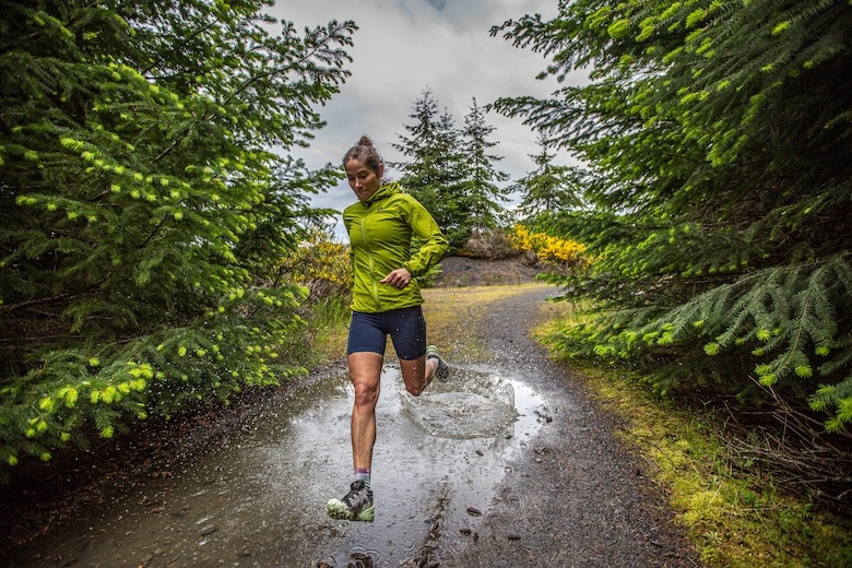 A woman runs through a muddy and wet dirt path in the forest. 