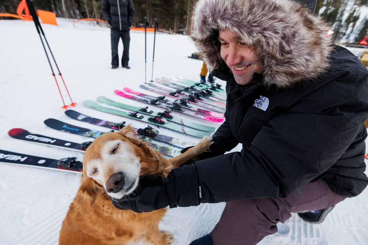 A man kneels down in the snow next to a bunch of skis to pet a dog