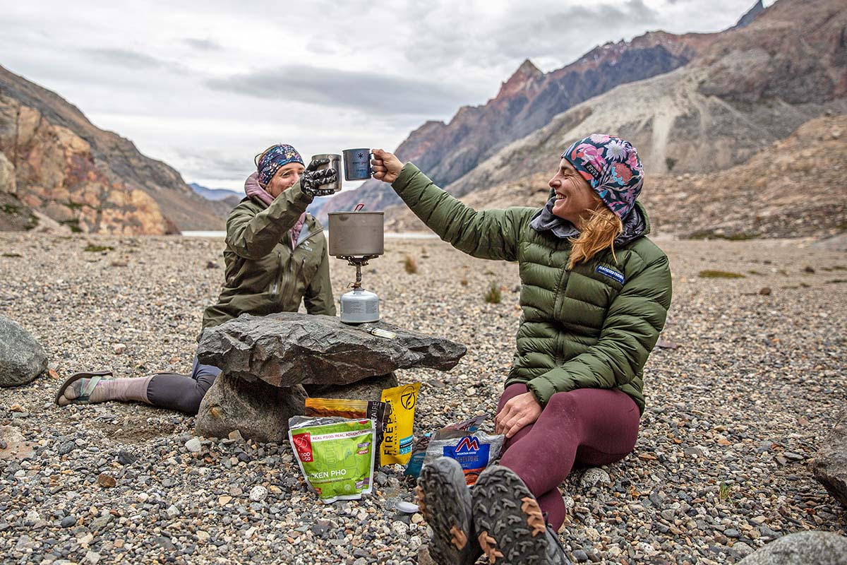 Two women smile and toast their drinks while preparing a meal with a backpacking stove