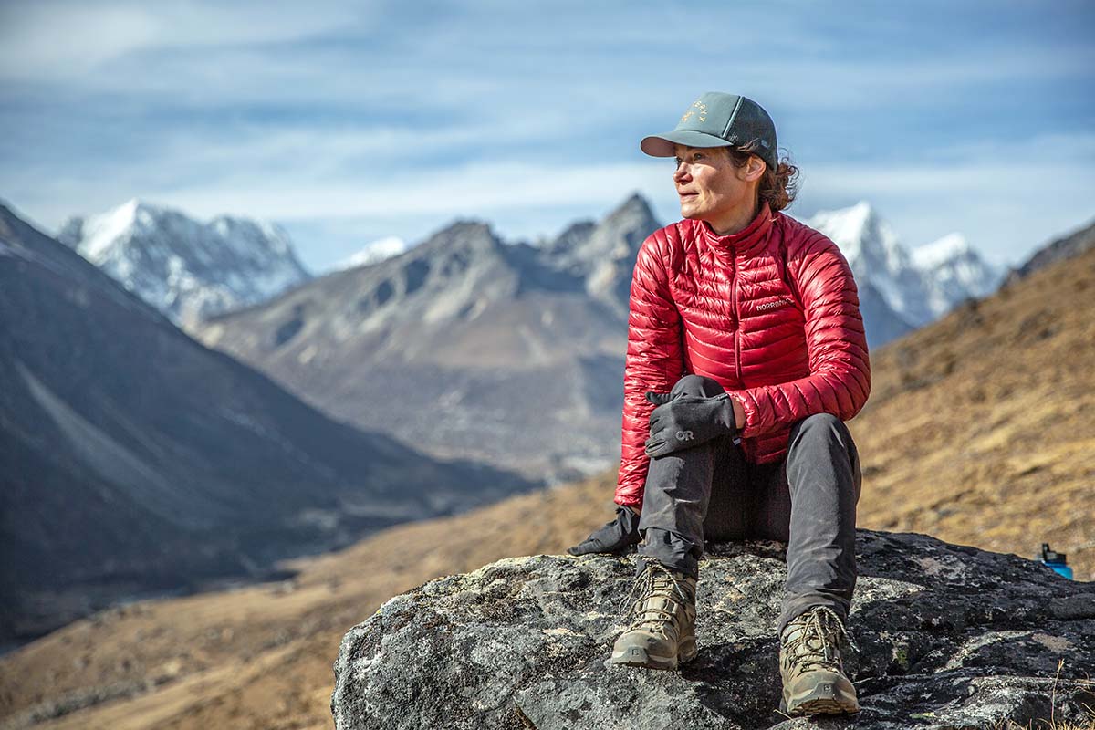 A woman sits in front of a mountain range in a down jacket