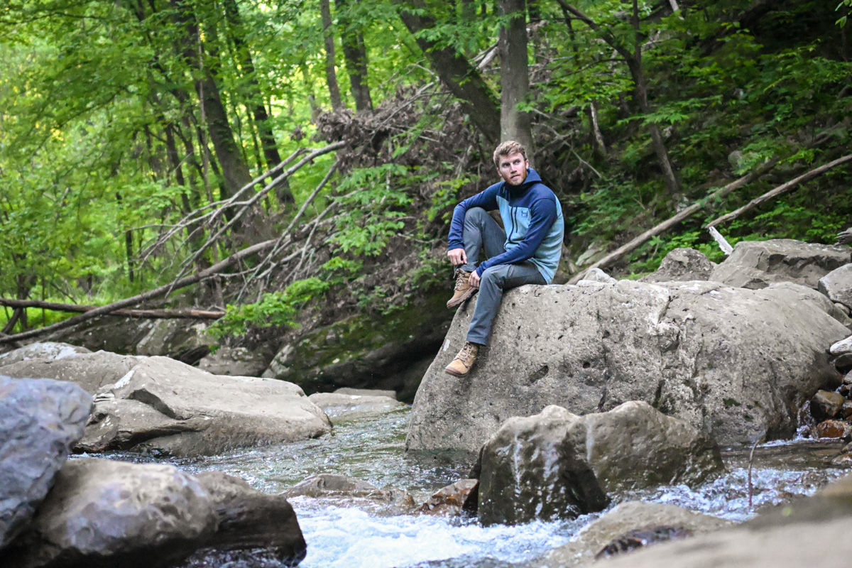 A man sits on a boulder wearing a fleece jacket near a river