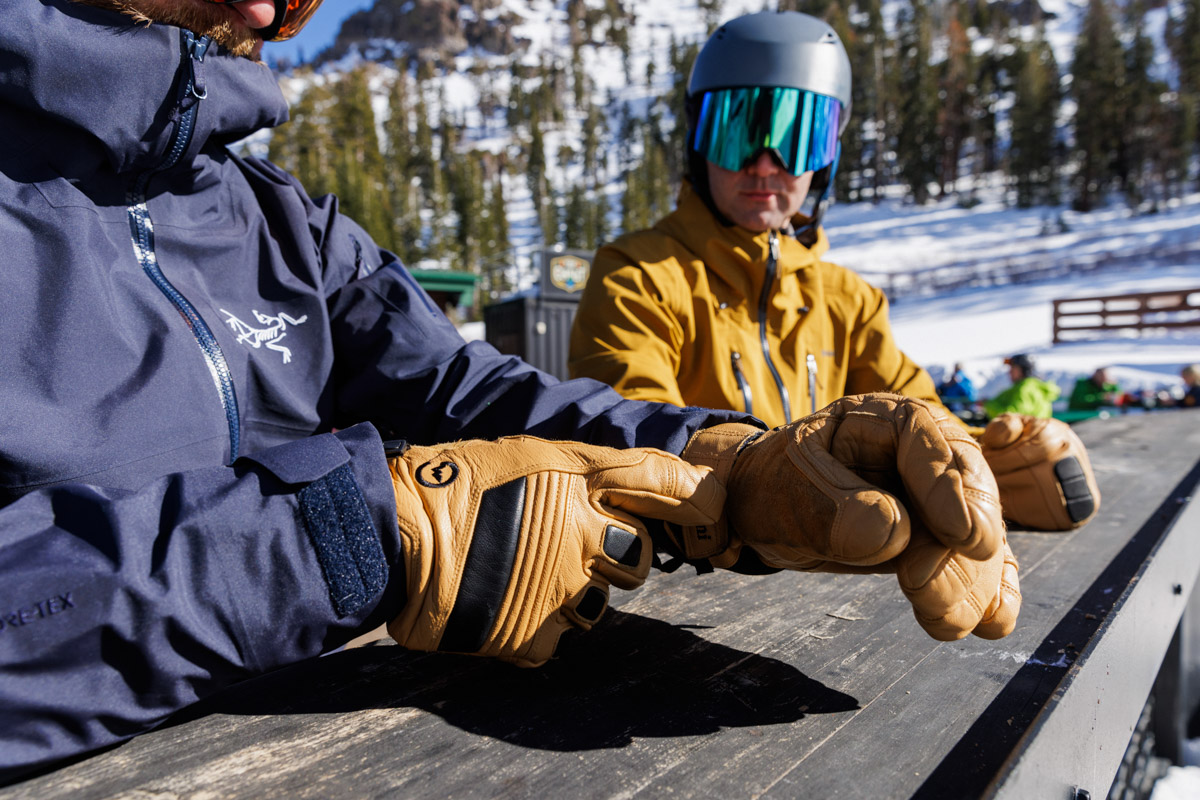 Two men sit at a picnic table at a ski resort, comparing their gloves