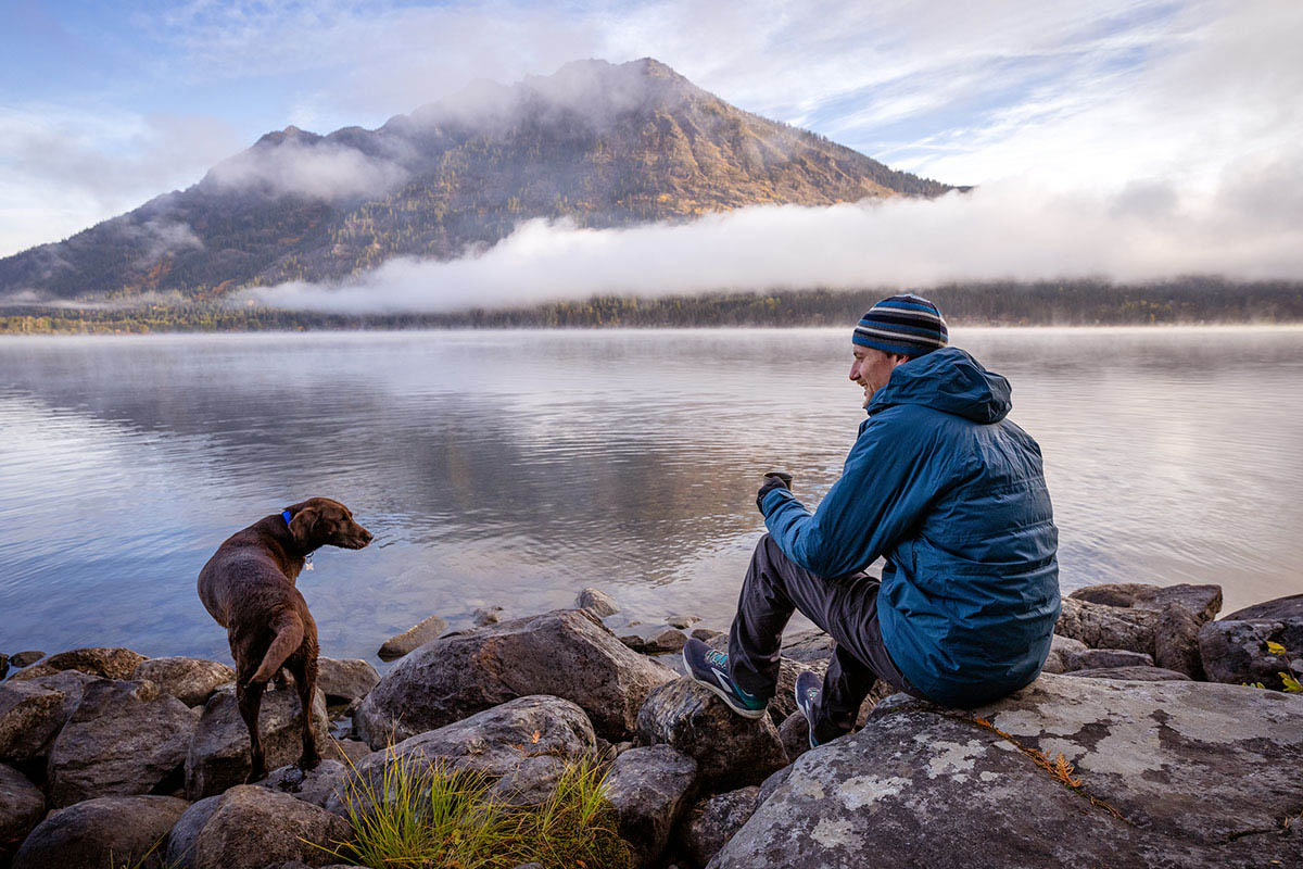 A man sitting by foggy lake with his dog while wearing the REI Stormhenge Down Hybrid