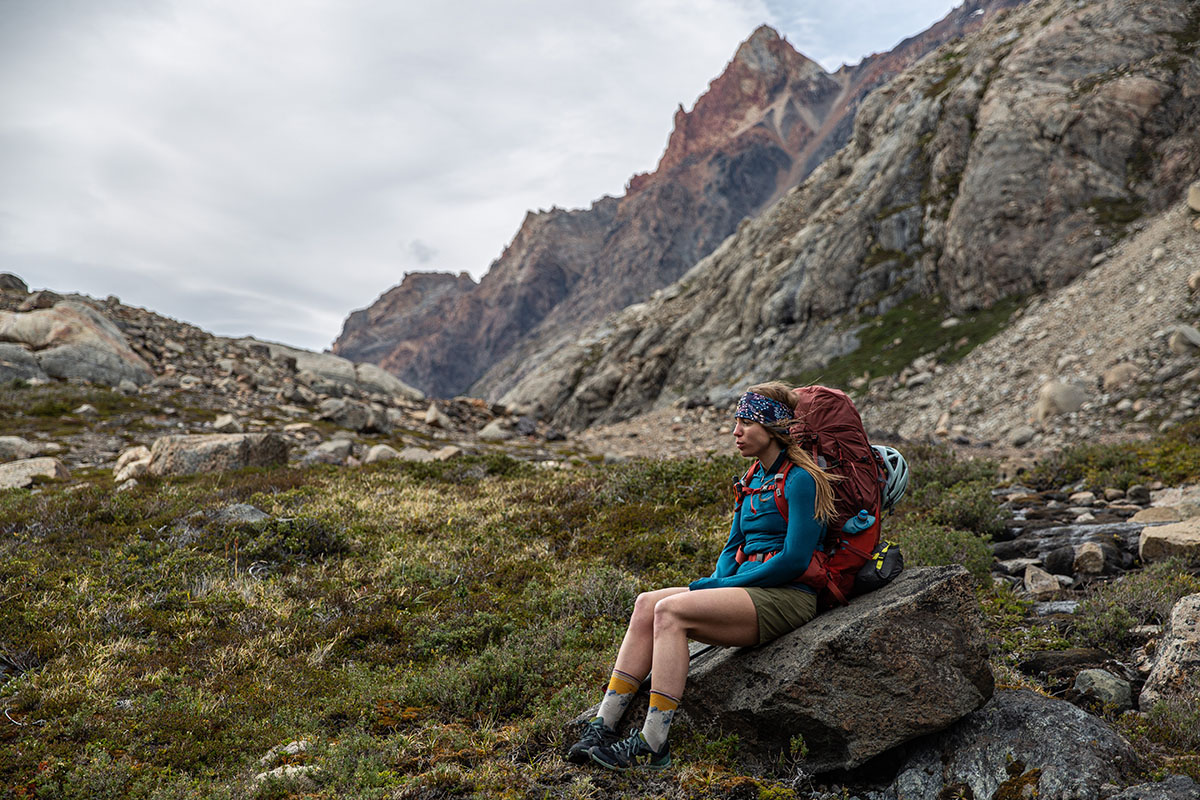 A woman wearing a merino base layer and a large backpack rests on a rock in the backcountry of Patagonia