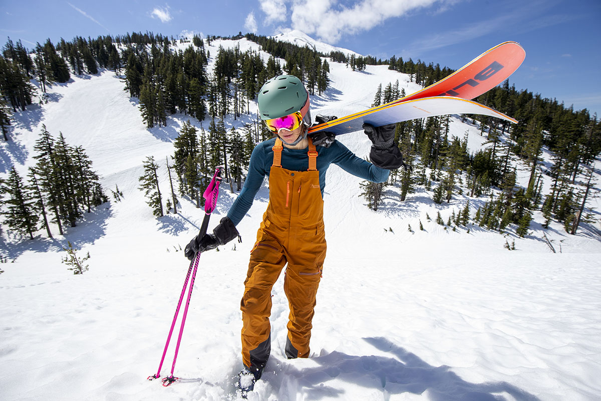 A woman in orange bibs bootpacks up a hill with her skis on her shoulder