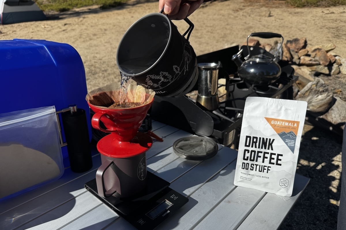 Water being poured from a Jetboil into a V60 dripper with a bag of coffee and a stove with a kettle and mokapot in the background
