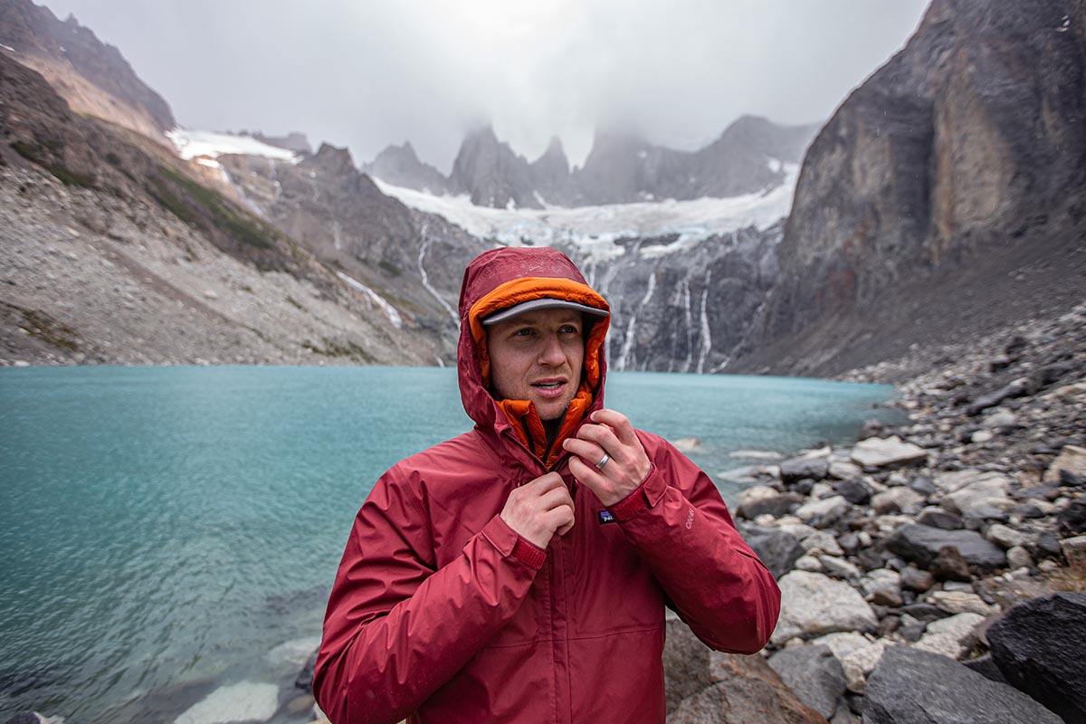 A man stands in front of a lake unzipping a jacket