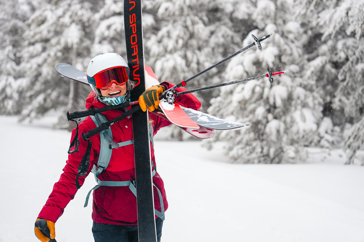 A woman carries skis in front of snow-covered trees