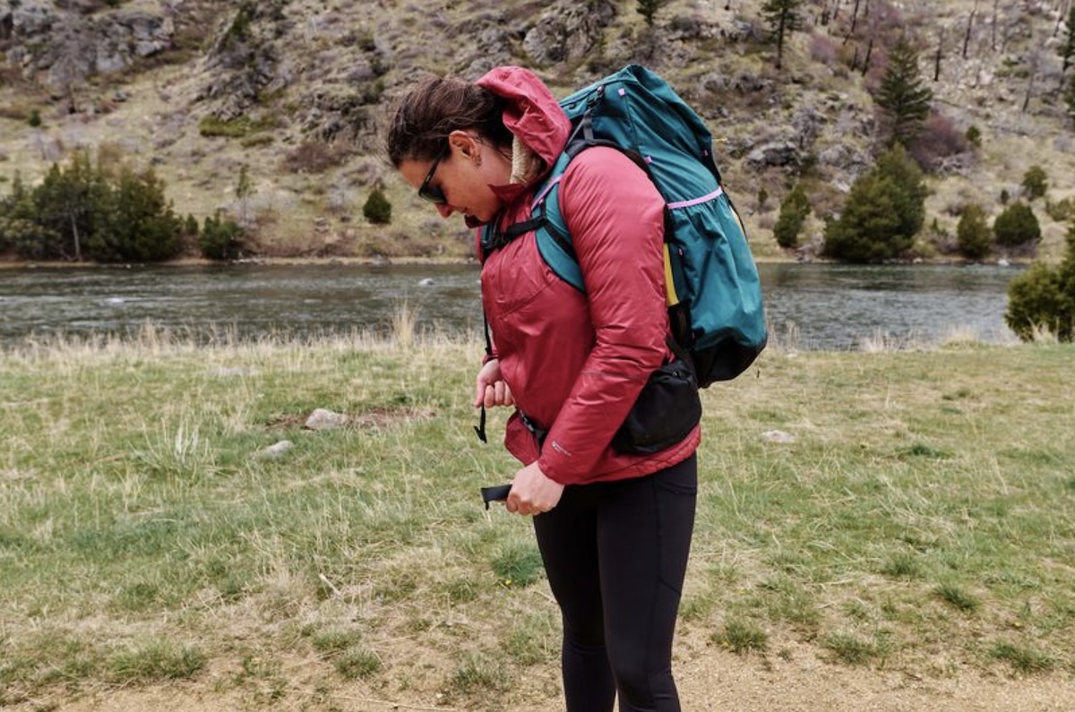 A woman stands next to a river in Montana adjusting the hip belt on the Mariposa backpack