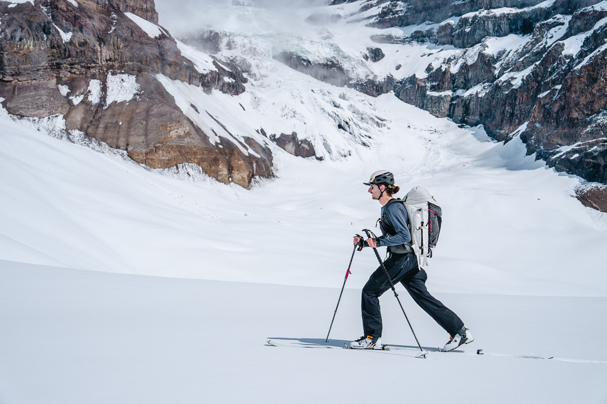 A man on skis skins up a hill with snowy mountain peaks in the background