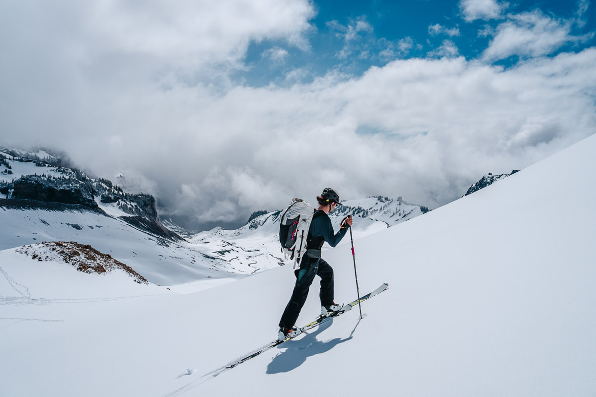 A man on skis skins uphill in fresh snow with mountains in the background