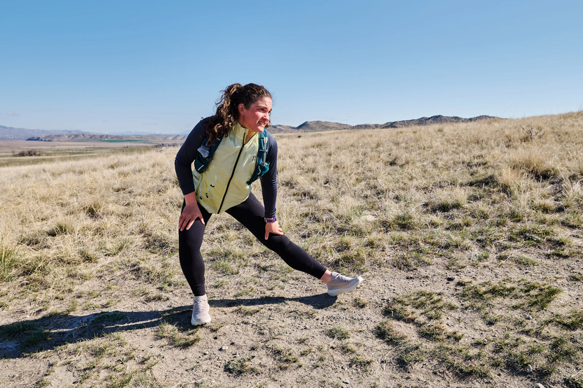 A woman wearing a running vest stops on a sunny trail to stretch