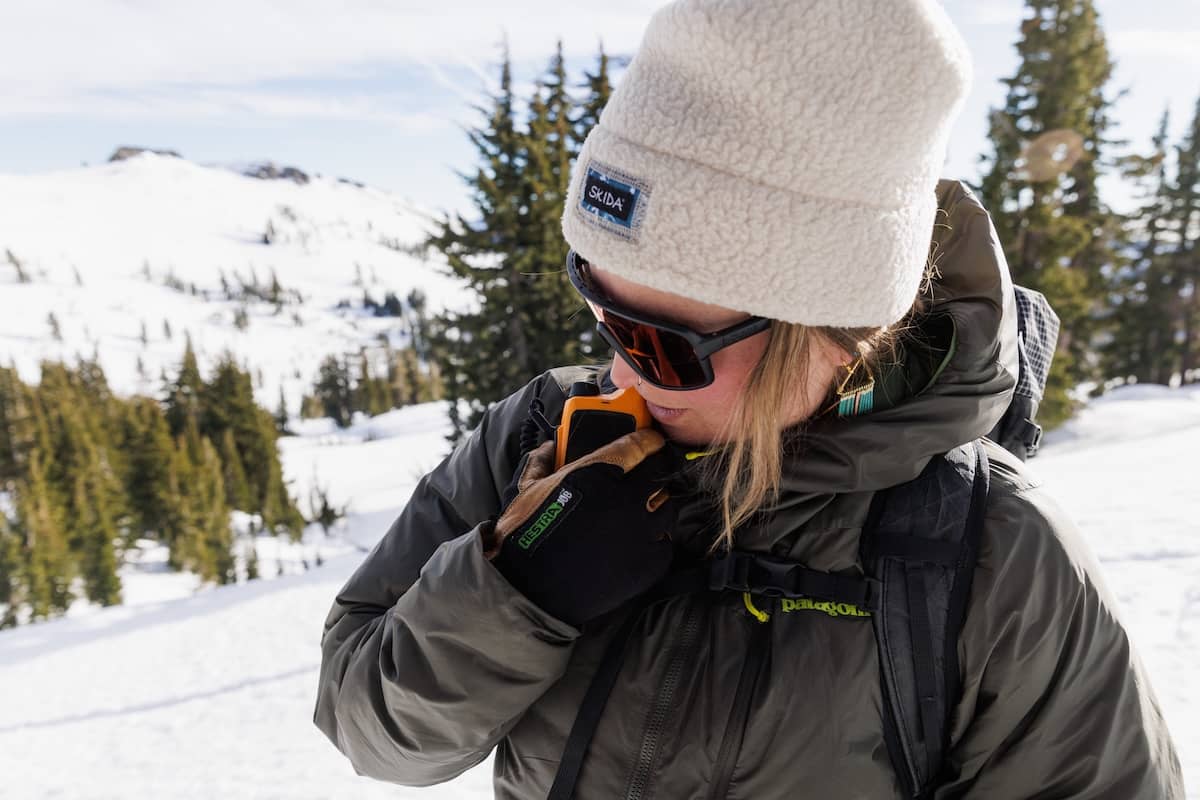 A woman talks into a walkie talkie in the snowy mountains while wearing the Patagonia DAS Light Hoody