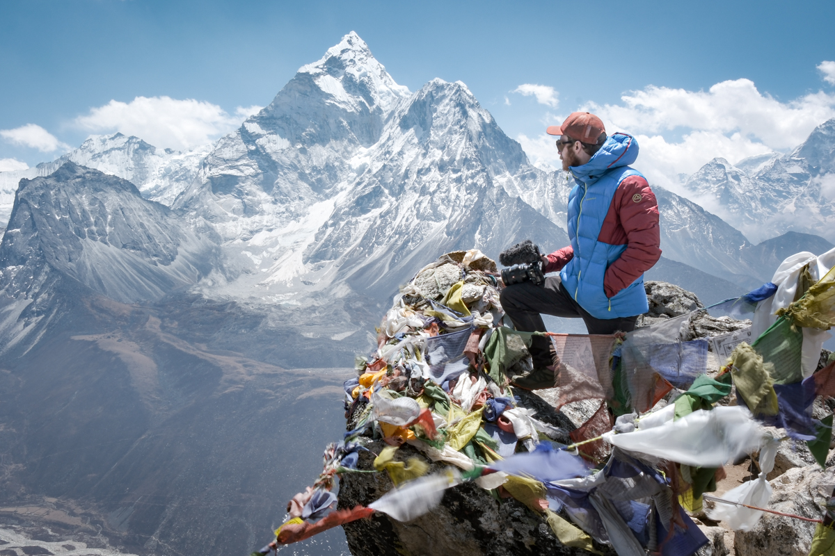 A man is standing on a peak in Nepal, with peace flags fluttering, looking at a view of Ama Dablam