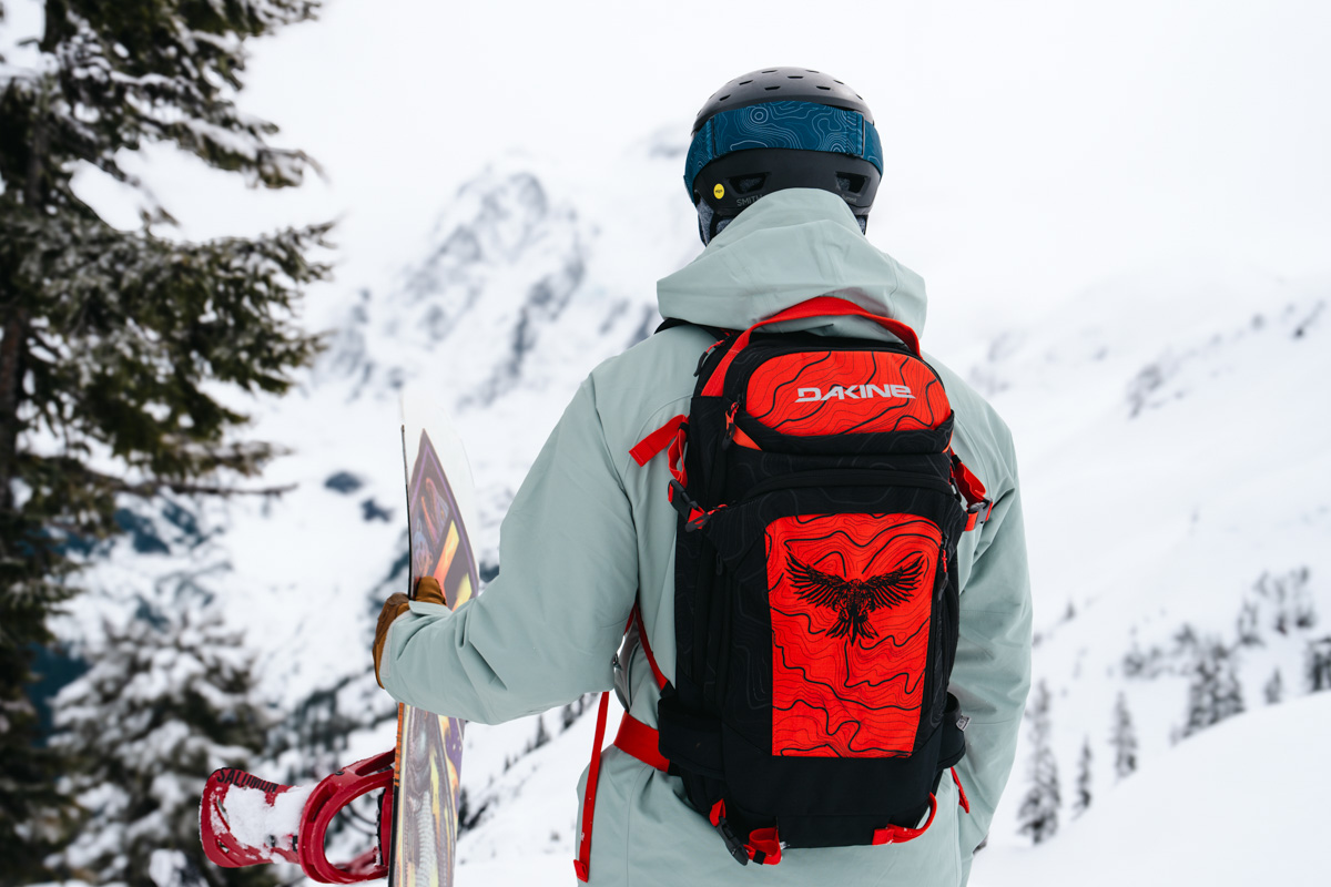 A man facing a snowy mountain background while holding a snowboard and wearing a red ski backpack
