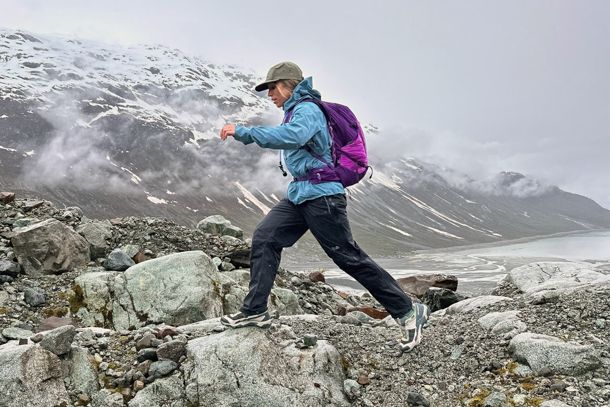 A woman stepping between rocks while hiking in a snowy Alaska landscape in a purple daypack