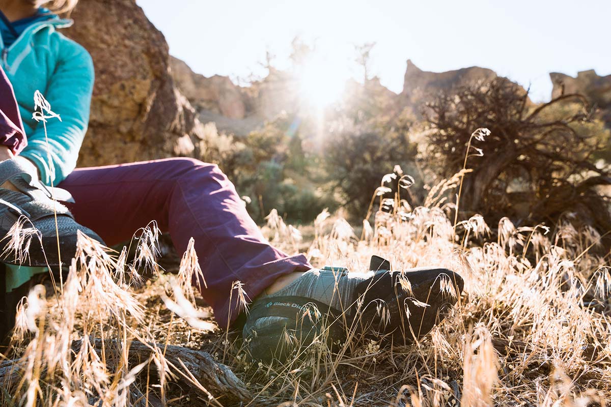 A woman puts on a climbing shoe while sitting on the ground in bright, late day sunlight