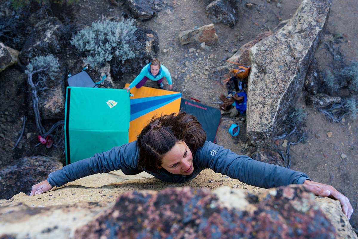 A woman climbs up a boulder with a crash pad and spotter below her
