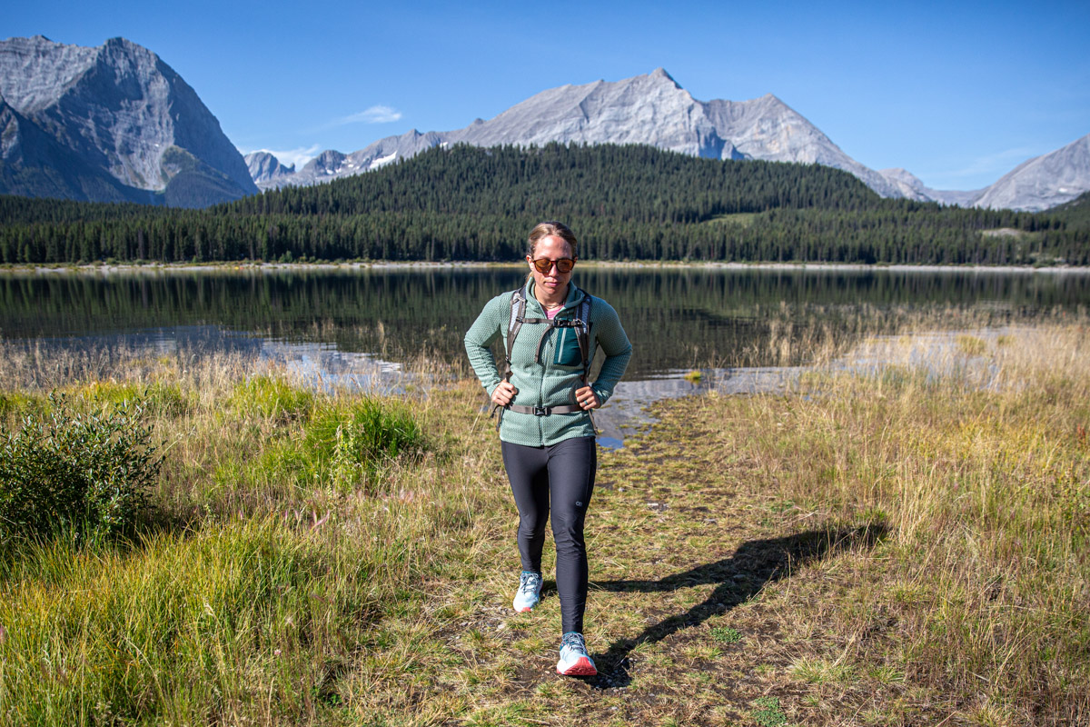 A woman in a fleece jacket walks toward us with a mountain range behind her