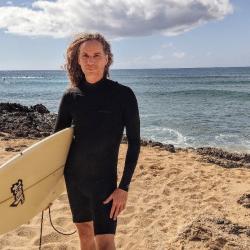 Michael standing next to the ocean wearing a wetsuit and holding a surfboard