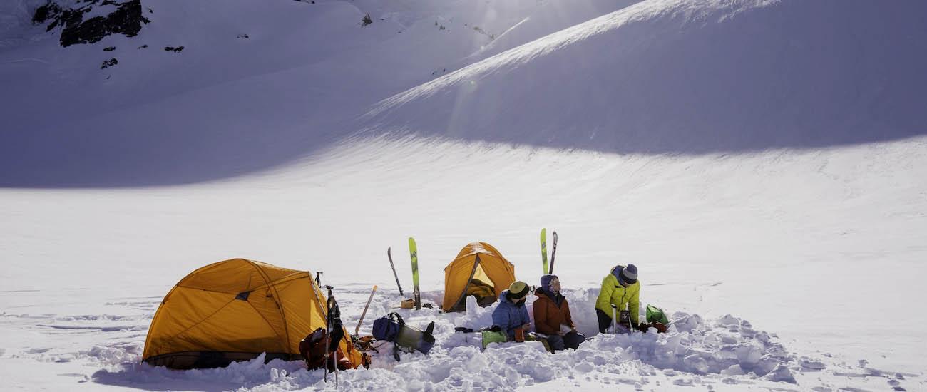 Three people sit in the snow next to tents and skis