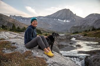 A woman sits on a rock in front of a mountain range with her dog